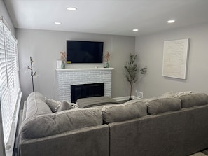 A modern living room with a fireplace, a large flat-screen TV, and a sectional sofa. - Deluxe 4 BR 2 BA Neighborhood Gem (Wilmington)