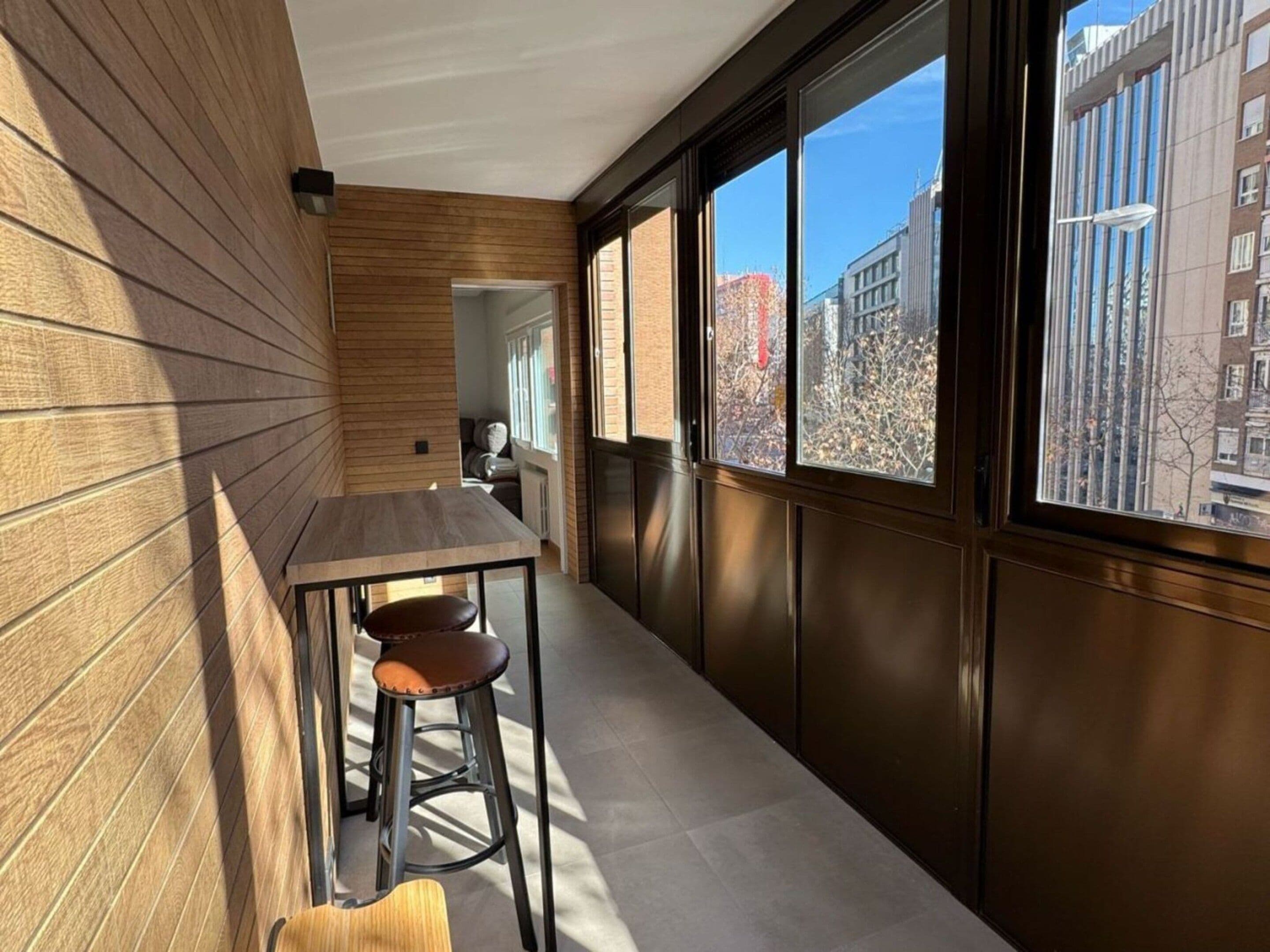 A narrow corridor with a wooden wall on one side and a window on the other, leading to a room with a table and stools.