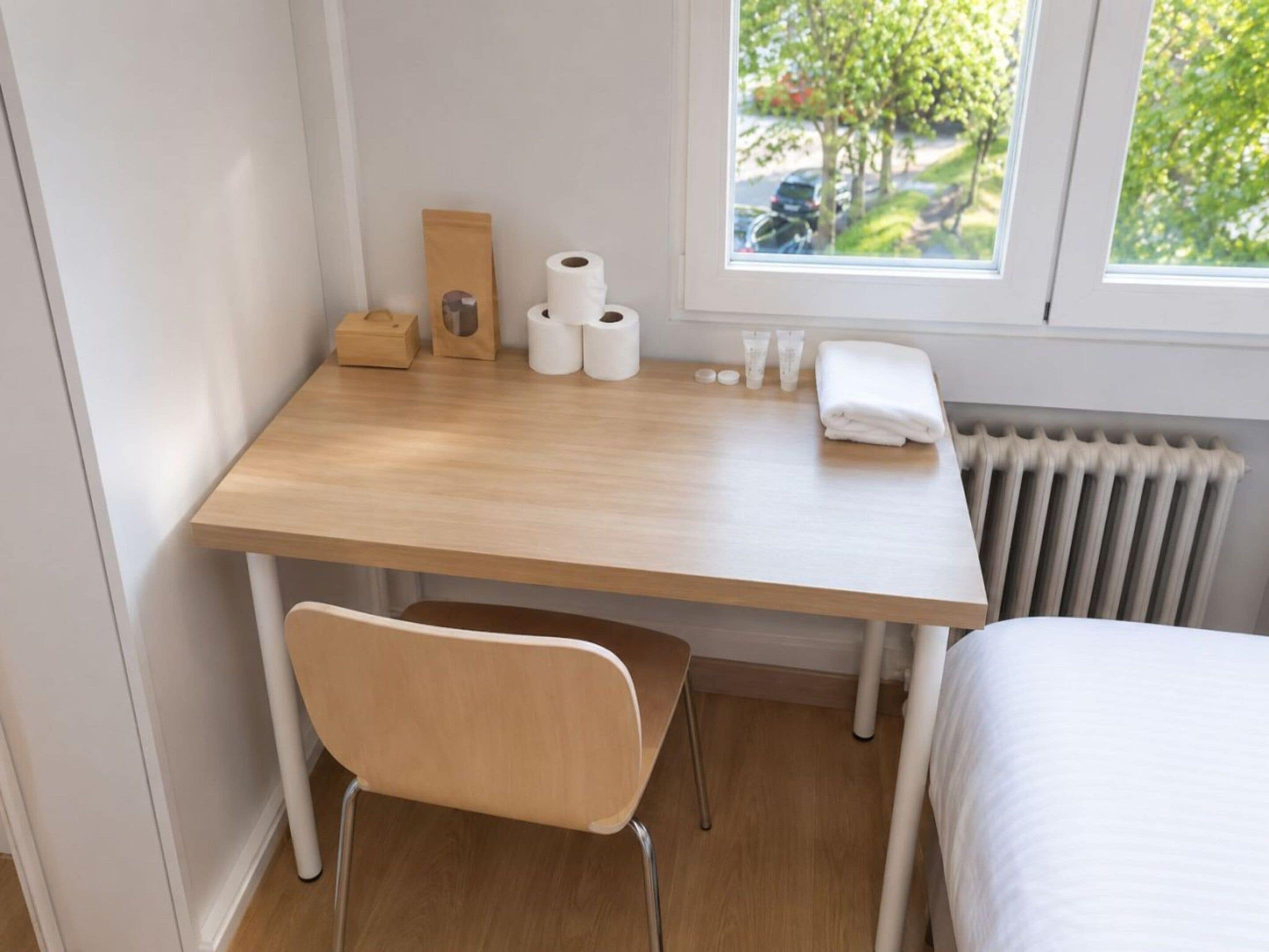 A small, neatly arranged room with a wooden desk, a chair, a radiator, and a window with a view of trees and parked cars.