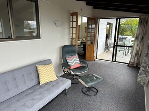 Ein Zimmer mit grauem Sofa, einem Drehsessel und einer Glastür zu einer Terrasse im Freien mit Tisch und Stühlen.