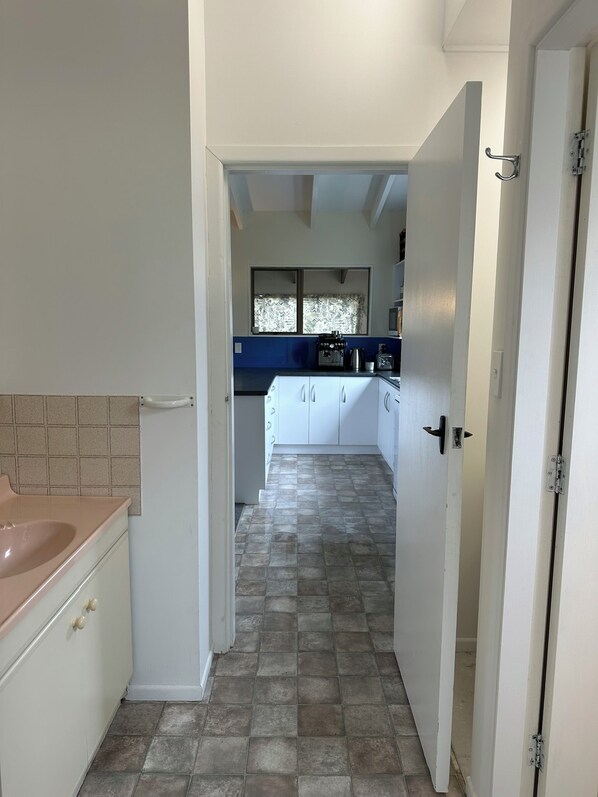 Een smalle gang die leidt naar een keuken met witte kasten, een blauw aanrecht en een raam met behang gordijnen.