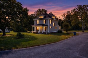 Ein zweistöckiges Haus mit Veranda, umgeben von Bäumen und einem grünen Rasen.