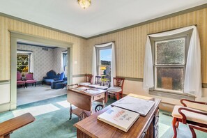 Quarto com escrivaninha de madeira, cadeiras, poltrona azul e uma grande janela com cortinas.