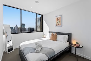 Een moderne hotelkamer met een groot raam, een bed met witte lakens, een nachtkastje met een lamp en een lijstwerk aan de muur.