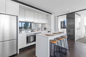 A modern kitchen with stainless steel appliances, a central island with stools, and a view of the cityscape through large windows. - Top Floor City Escape | 180° Views, Pool, Parking (Melbourne)
