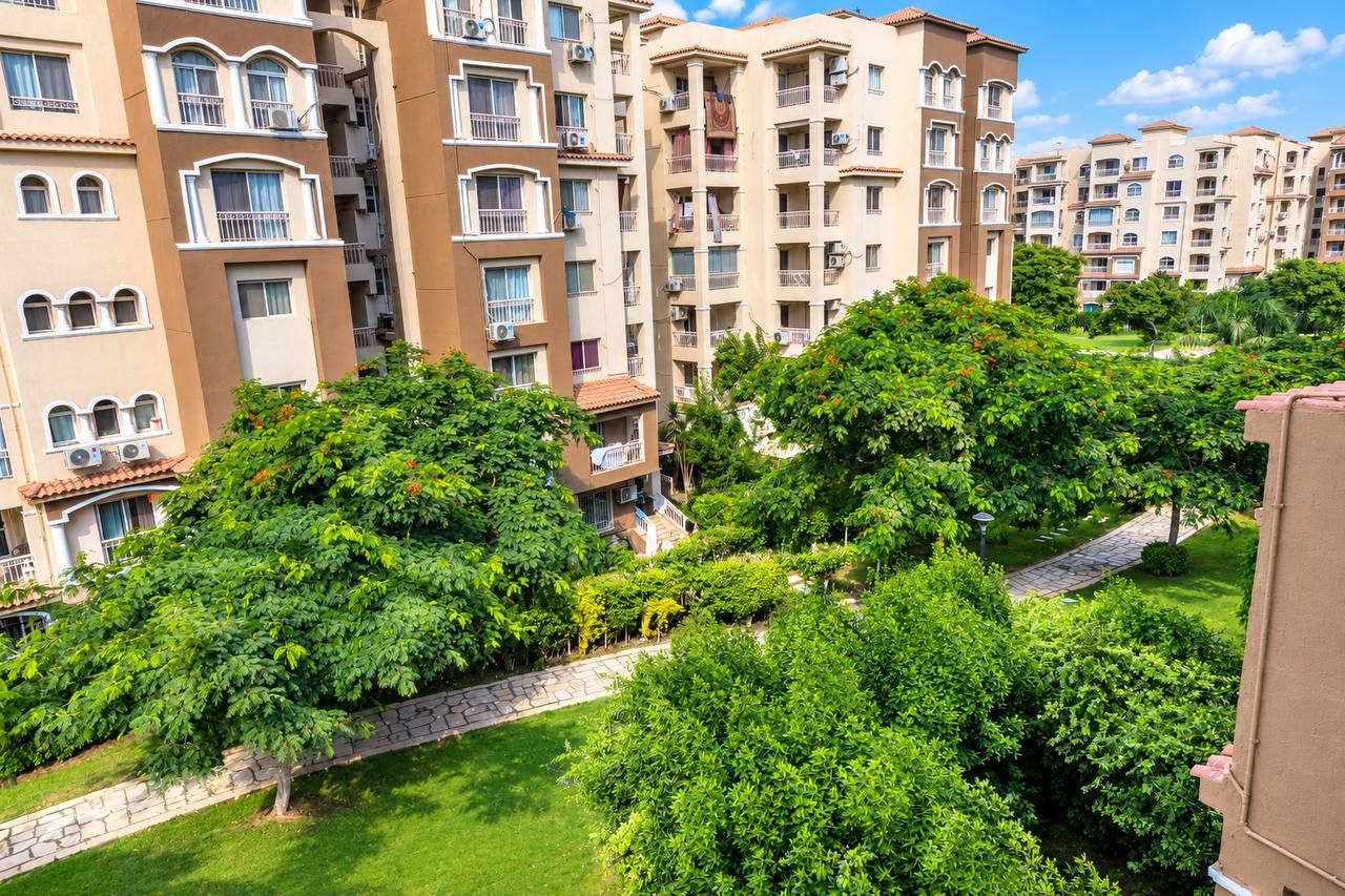 Un immeuble résidentiel à plusieurs étages, doté de balcons, entouré d’une végétation luxuriante et bordé d’un chemin pavé de pierres.