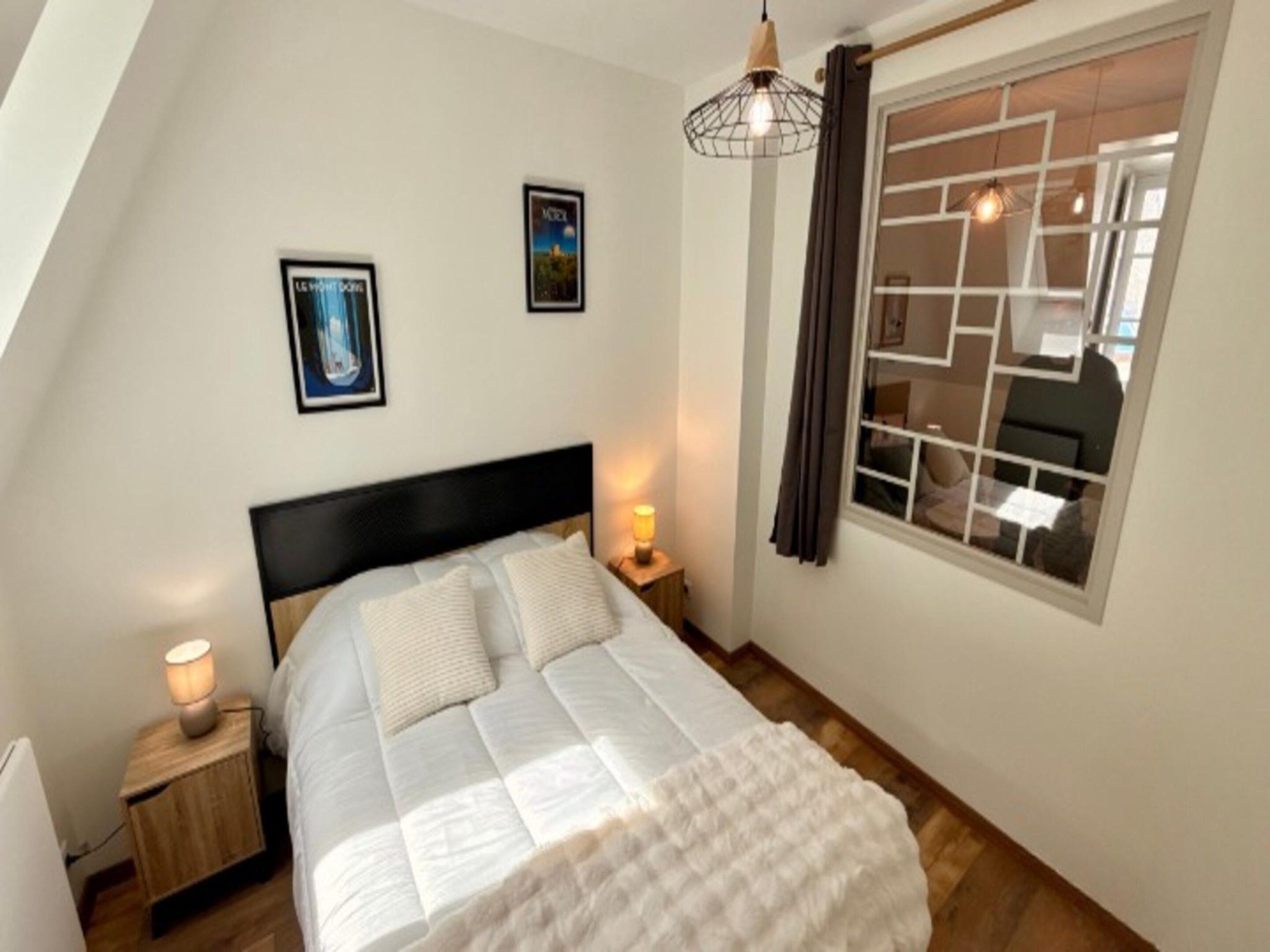 A bedroom with a bed, a nightstand, a wall-mounted lamp, a window with curtains, and two framed pictures on the wall.