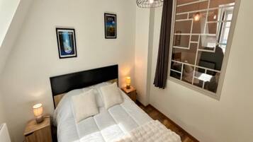 A bedroom with a bed, a nightstand, a wall-mounted lamp, a window with curtains, and two framed pictures on the wall.