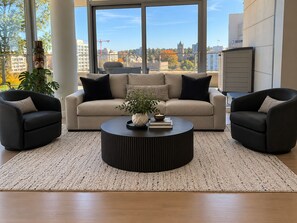Una sala de estar moderna con un sofá color beige, dos sillones negros, una mesa de centro negra redonda y vistas de un paisaje urbano a través de grandes ventanales.