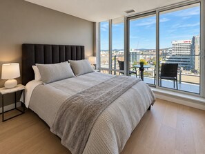 Camera da letto moderna con un letto grande, un comodino, una lampada e vista sulla città attraverso ampie finestre.