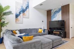 A modern living room with a grey sectional sofa, a flat-screen TV, and abstract wall art. - 7BR Villa Near Disney / FREE Pool Heat, Hot Tub, Game Room / Sleeps 14 (Clermont)