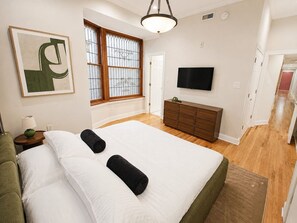A hotel room with a large bed, a TV, a dresser, and a window with bars. - Timeless Elegance on RQ | By Rafa Properties (Philadelphia)