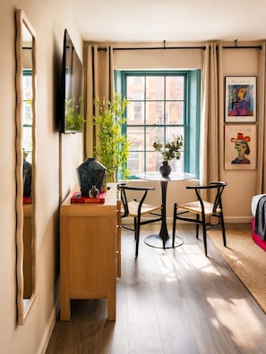 Een kamer met een houten bureau, twee stoelen, een aan de muur bevestigde televisie, een raam met gordijnen en kunstwerken aan de wand.