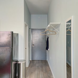 A narrow hallway with a metallic refrigerator on the left, a clothes hanger with a garment on the right, and a closed door at the end. - Charming Center City Studio | By Rafa Properties (Philadelphia)