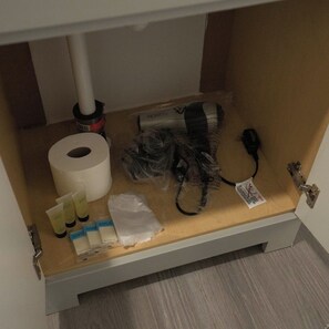 Een klein opbergvak onder een kastje, met een rol toiletpapier, een zwart voorwerp, een witte plastic zak en enkele kleine flesjes.