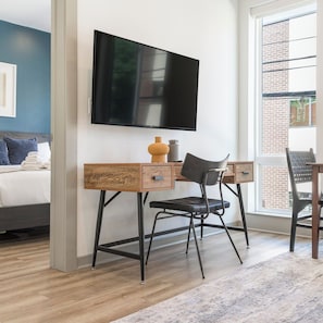 A modern hotel room with a wooden desk, a flat-screen TV, a bed with blue pillows, and a view of a brick building through a large window. - Comfortable 1BD near UPenn & Drexel | Queen Bed (Philadelphia)
