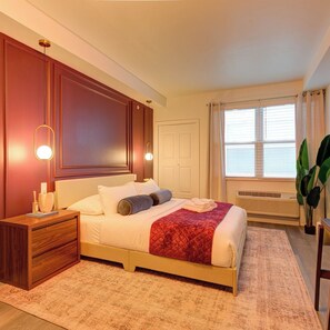 Quarto de hotel com cama, mesa de cabeceira, luminária e janela com cortinas.