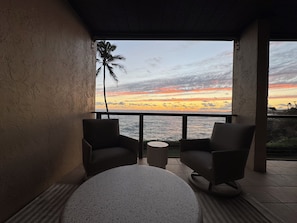 Een balkon met rieten stoelen en een ronde tafel, met uitzicht op een zonsondergang boven de oceaan.
