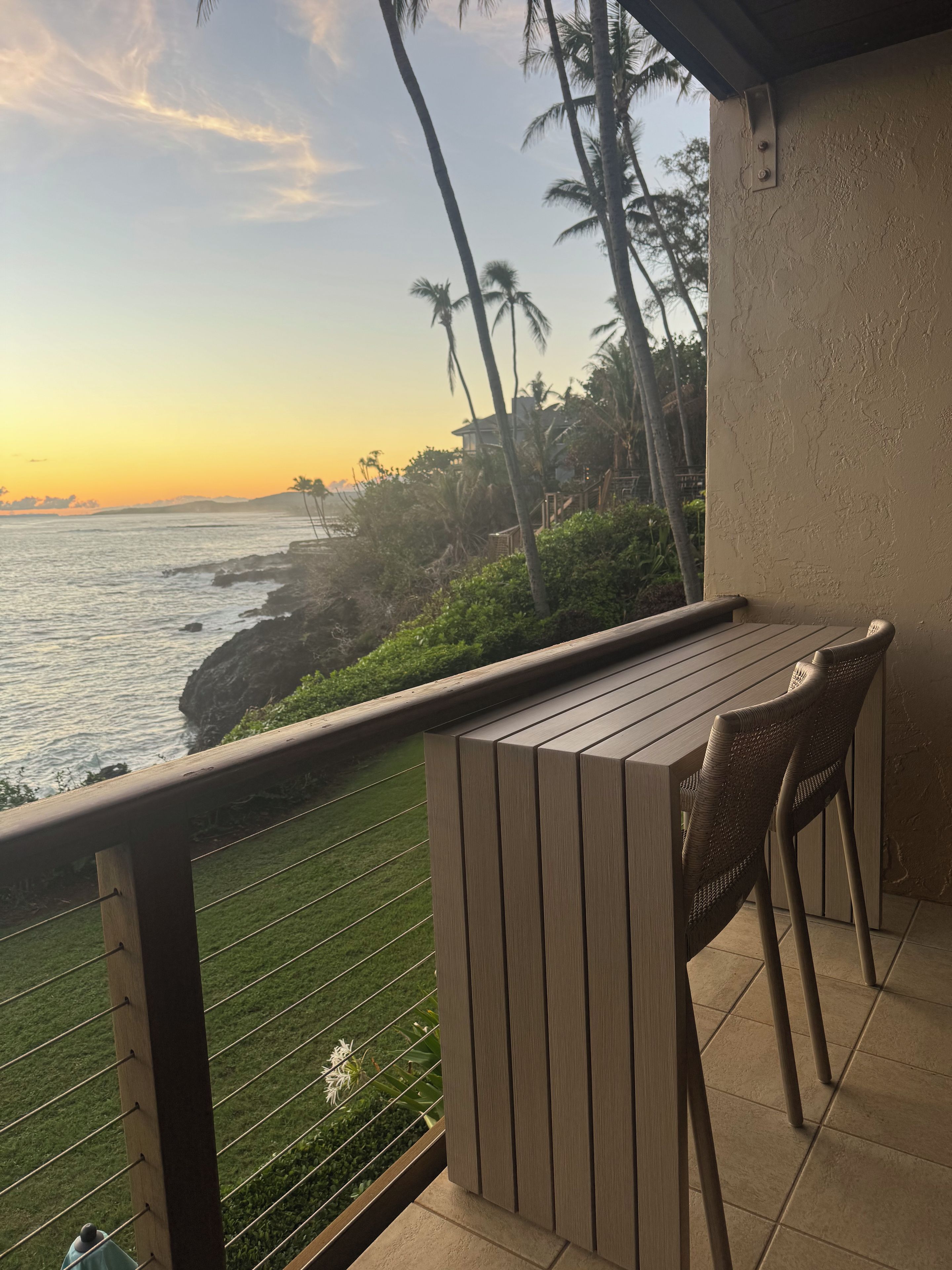 A balcony with a wooden table and chairs overlooking a coastal area with palm trees and a view of the ocean.