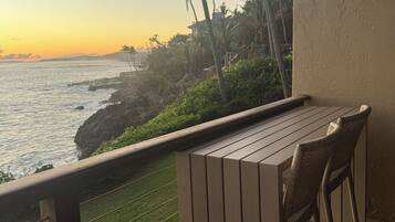 A balcony with a wooden table and chairs overlooking a coastal area with palm trees and a view of the ocean.