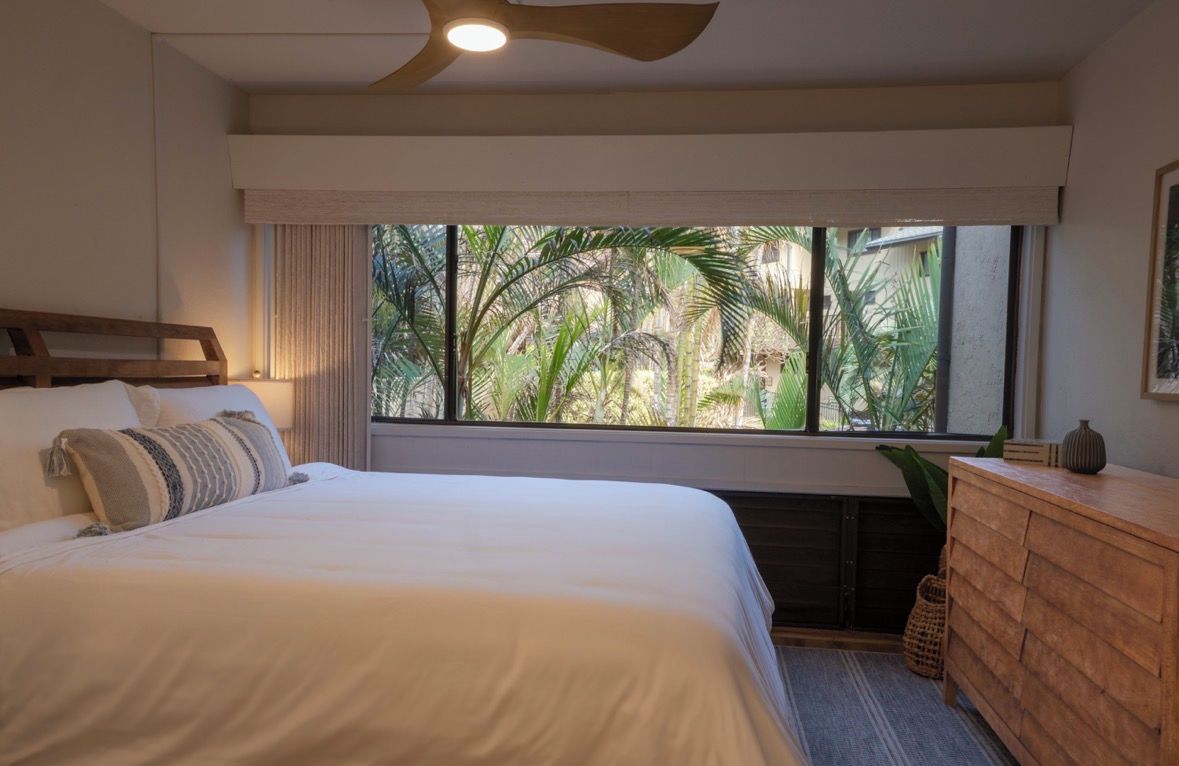 Une chambre d’hôtel avec un grand lit, une tête de lit en bois, une fenêtre donnant sur des palmiers, un ventilateur de plafond et une commode en bois.
