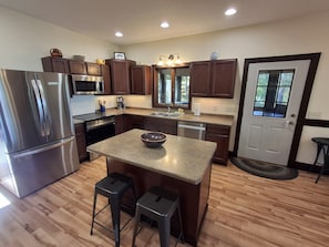 A modern kitchen with a stainless steel refrigerator, wooden cabinets, a central island with stools, and a window providing natural light. - Spacious Lakehouse on Lac Courte Oreilles (Stone Lake)
