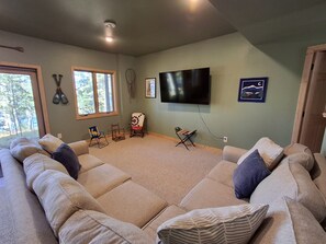 A living room with a large sectional sofa, a flat-screen TV mounted on the wall, and a window with a view of trees. - Spacious Lakehouse on Lac Courte Oreilles (Stone Lake)