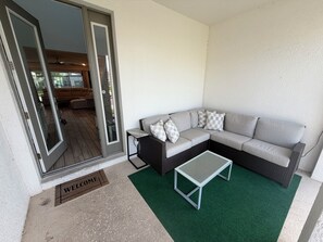 Un espacio de estar al aire libre cubierto con un sofá y una pequeña mesa, una puerta que da a un patio, y un felpudo.