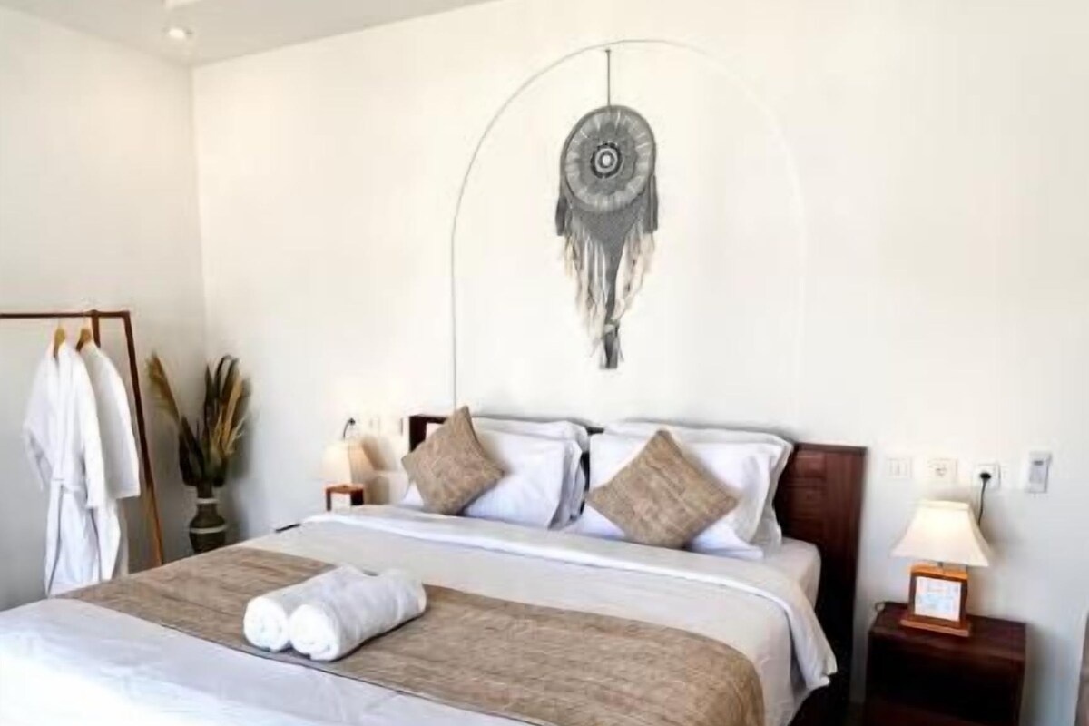 Een opgemaakt bed met houten frame, een nachtkastje met een lamp en een decoratief object aan de muur.