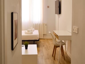 Ein Hotelzimmer mit Bett, Schreibtisch, Stuhl, einem kleinen Tisch mit Pflanze und Fernseher an der Wand.