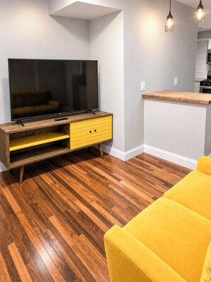 Una sala moderna con un televisor de pantalla plana sobre un mueble de madera, un sofá amarillo y una zona de cocina con encimera de madera.