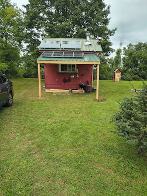 A small red wooden structure with a green roof, a barbecue grill, and a car parked nearby. - Beautiful cabin in soothing Livonia perfect for a relaxing getaway. Off grid . (Livonia)
