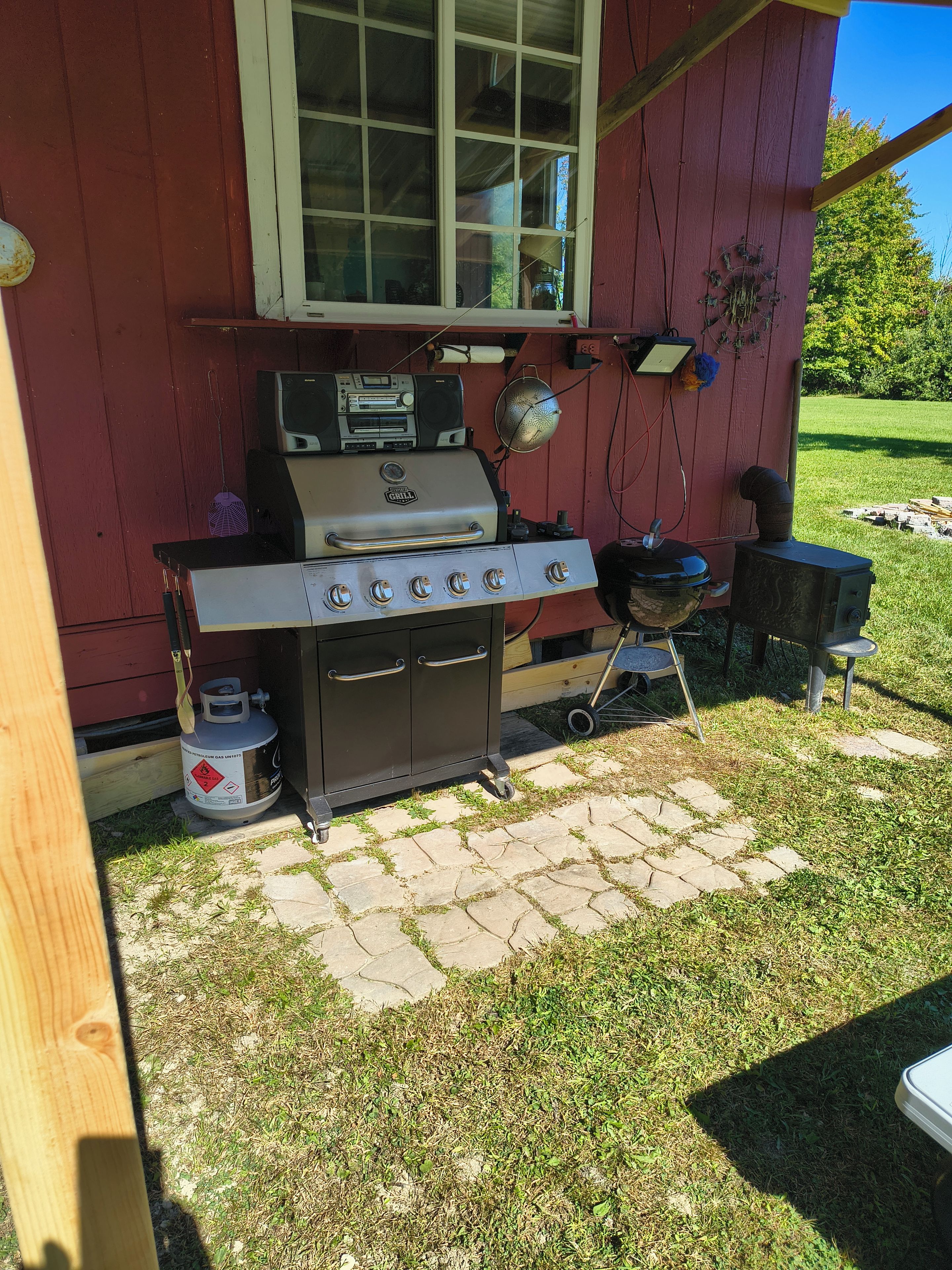 A portable gas grill with a side burner and a built-in oven, positioned on a stone patio outside a red building.