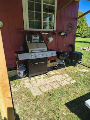 A portable gas grill with a side burner and a built-in oven, positioned on a stone patio outside a red building.