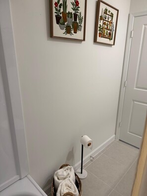 A hallway with a white door, a framed plant illustration on the wall, a floor lamp, and a basket with a towel. - Golden Pine Haven, Luxury 5-Bedroom Vacation Home. (Homestead)