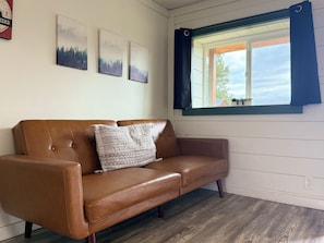 A brown leather sofa with a patterned pillow, a window with blue curtains, and three landscape paintings on the wall. - Peaceful Mountain Retreat Near Durango with Beautiful Nature Views (Bayfield)
