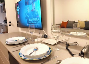 Uma área de refeições com uma televisão exibindo um filme, mesa posta com pratos, copos e talheres, e um sofá com almofadas ao fundo.