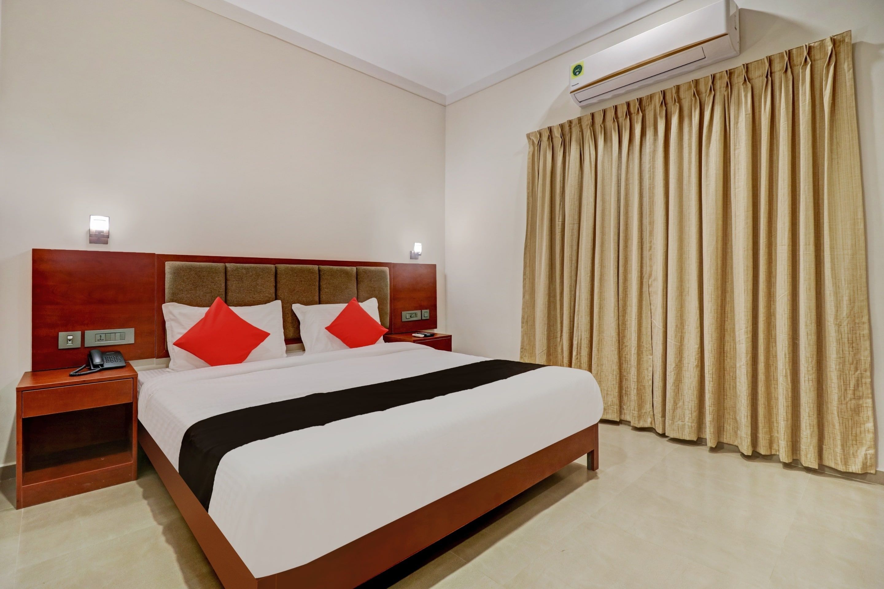 A hotel room with a bed, a nightstand, a telephone, an air conditioning unit, and a window with curtains.