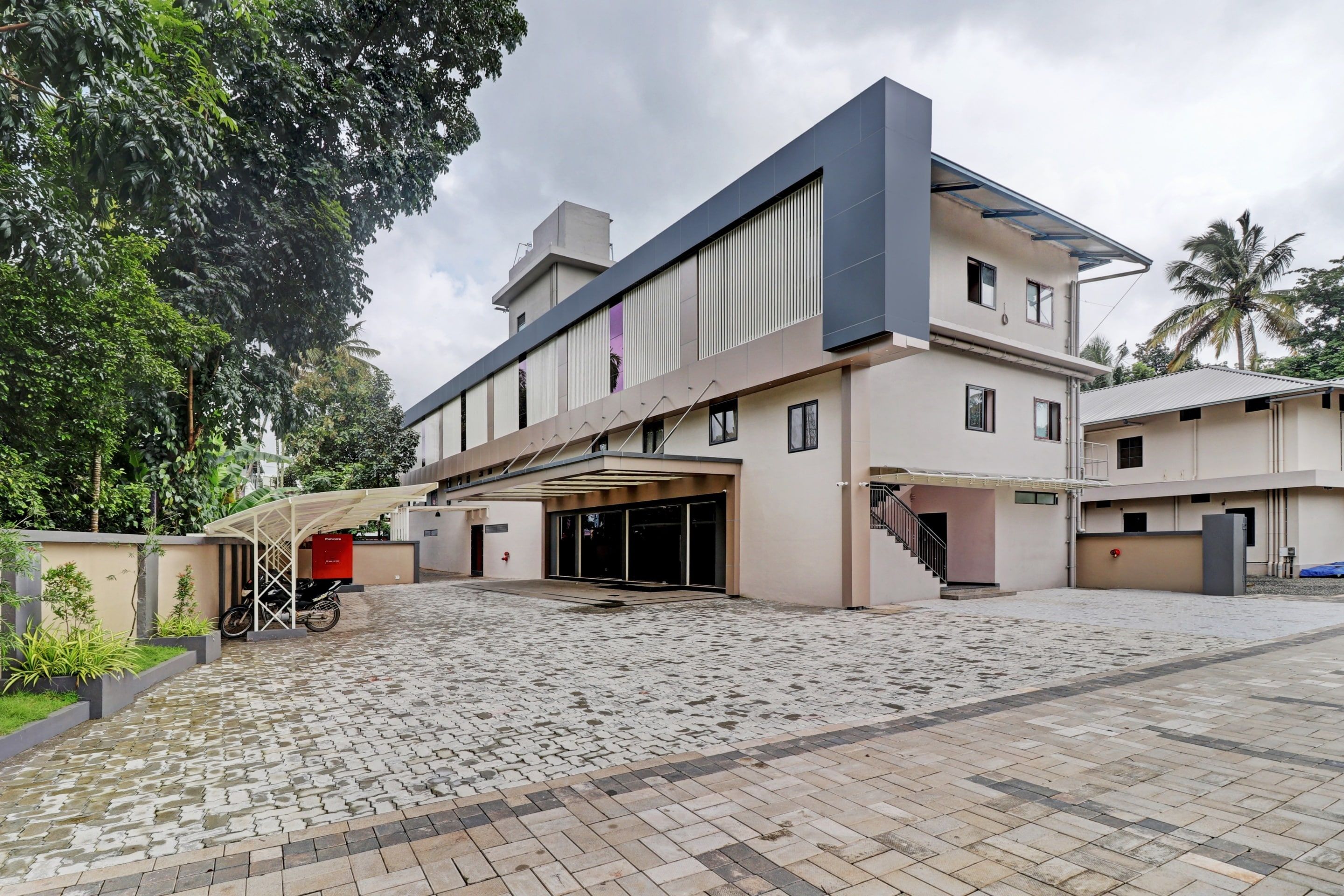 Un edificio moderno de varios pisos con un acceso cubierto, patio empedrado y arbolado.