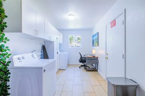 A laundry room with a washing machine, dryer, and ironing board. There is a desk with a chair and a lamp. A window provides natural light, and an exit sign is visible. - Family Retreat /Heated Pool/Billard/FirePit 12pp (Hollywood)