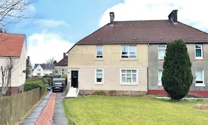 A two-story residential building with a red-tiled roof, a front door, and a small front yard with a trimmed bush. - Spacious, quiet 2 bedroom garden flat sleeps 4 (Coatbridge)