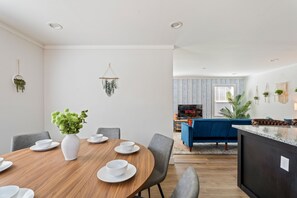 A modern living space with a dining area featuring a wooden table and chairs, a blue sofa, and a TV in the background. - 13 mins to Campbell University Boho Retreat (Angier)