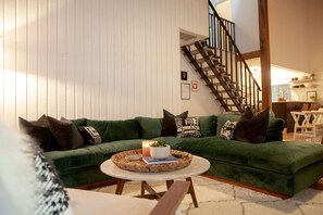Ein grünes Sofa mit dekorativen Kissen, ein runder Tisch mit Kerze und Pflanze sowie eine Treppe im Hintergrund.