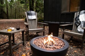 A fire pit with logs, two chairs with plaid blankets, a table with drinks, and a forested background. - Hood Hideaway: Dog-Friendly Mt. Hood Cabin, Private Hot Tub, Sleeps 8 (Rhododendron)