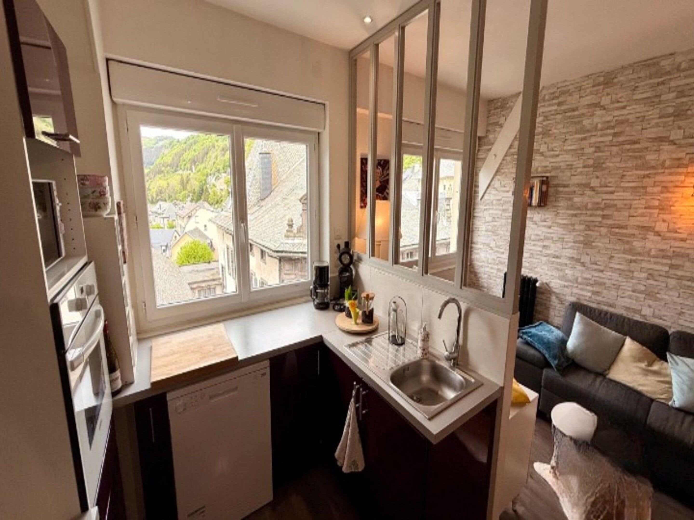 A compact kitchen with a large window, a built-in dishwasher, and a stone wall.