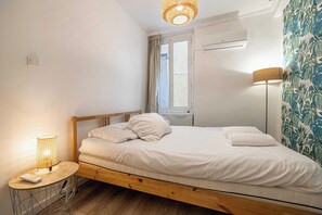 Ein Schlafzimmer mit einem Holzbett, einem kleinen Nachttisch mit Lampe, einem Fenster mit Vorhängen und einer wandmontierten Klimaanlage.
