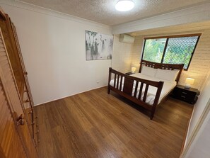A bedroom with a wooden bed, a nightstand, a wall-mounted air conditioner, and a window with a view of greenery. - Manly Bayside Retreat 3BR 2BA Free Parking (Manly)