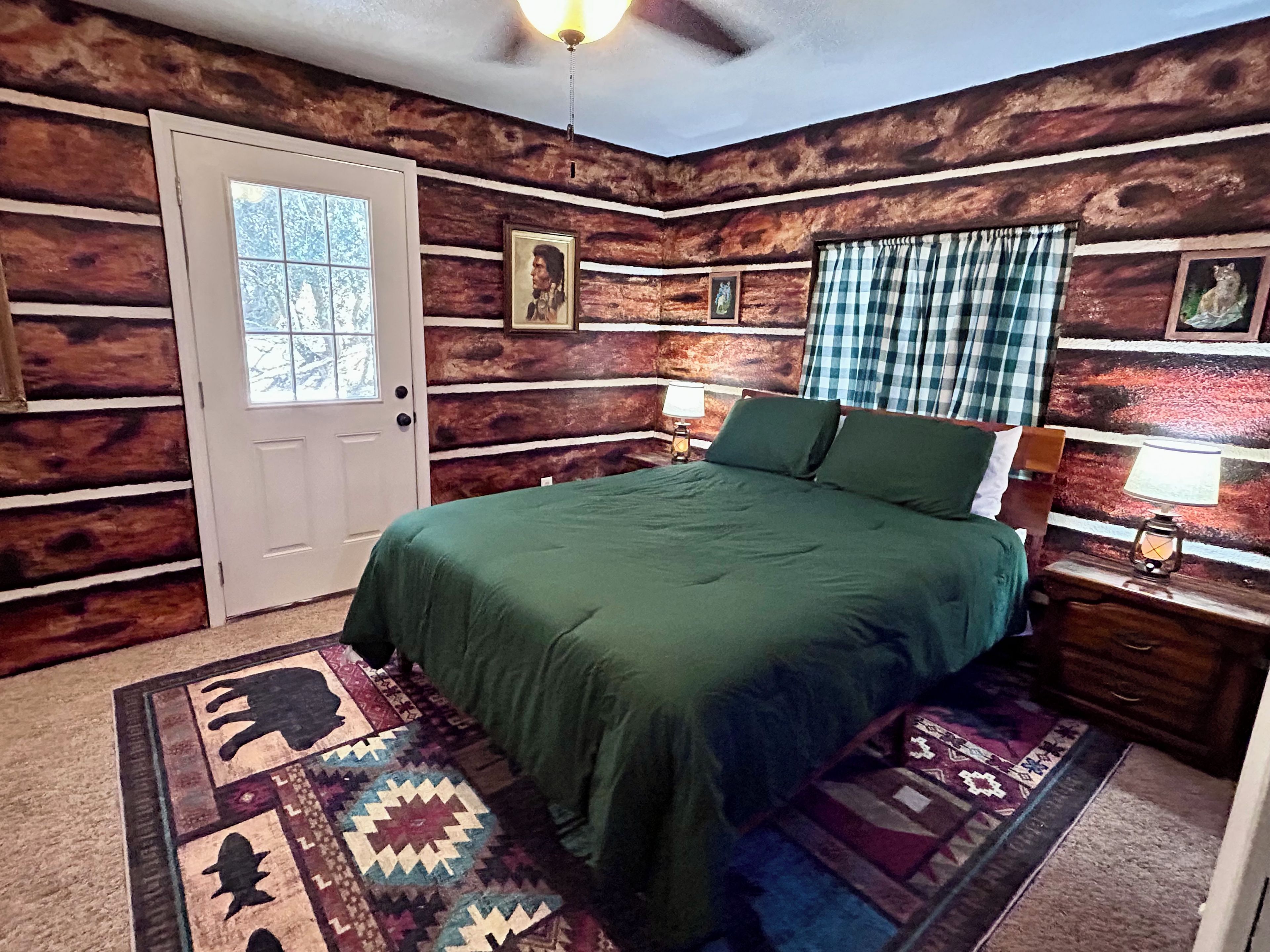 A bedroom with a wooden log wall, a bed with green bedding, a window with curtains, and a rug with a geometric pattern.