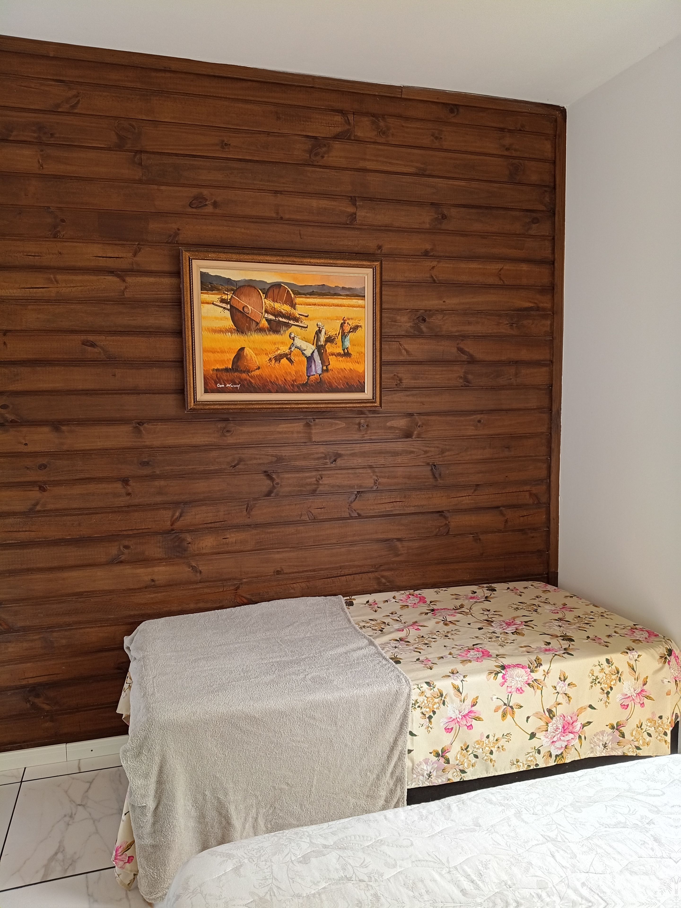 Une pièce avec un mur en bois, un lit recouvert d’une courtepointe à motifs floraux et un tableau encadré représentant un paysage rural.