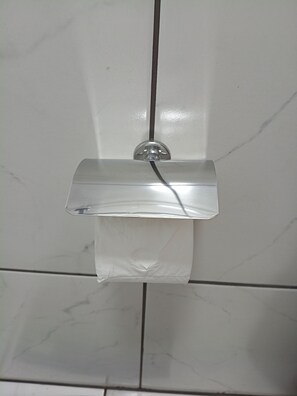 Un baño con pared de azulejos blancos, un jabonera de vidrio transparente fijada a la pared y un rollo de papel higiénico colgando debajo.